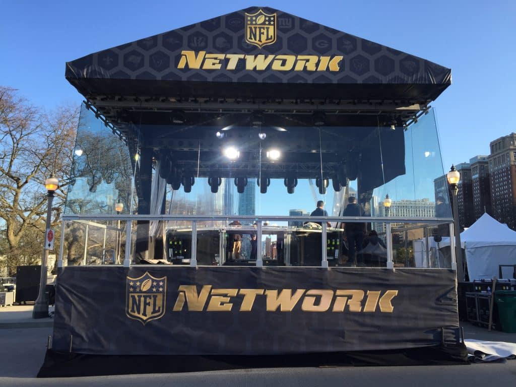 Looking at the branded tech area at an NFL NEtwork event. The tech area is raised on a low stage and surrounded by soft goods and topped with a roof. The roof and understructure aare branded with black soft goods tat have the NFL logo next to the work "network" , both in gold color. The middle section, between the deck and roof, is a clear plastic to the techs can see out, while still being protected from the elements.