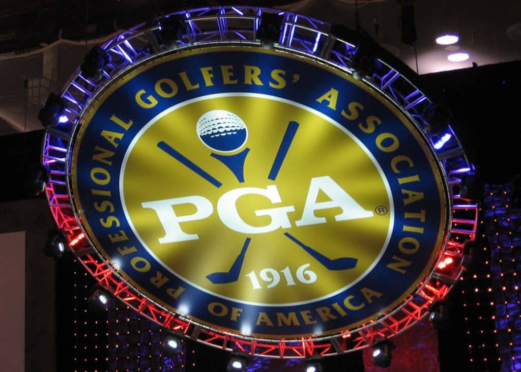 round scoft good. the center is yellow with the word PGA in white. 2 golf clubs cross behind the words. Outside the yellow center circle is a dark blue circular border with yellow words that say "professional golfers' association of america". Then outside of that is another circular border where the top is purple and the bottom is red. It hangs at an angle above head height and the background of the venue is just dark.