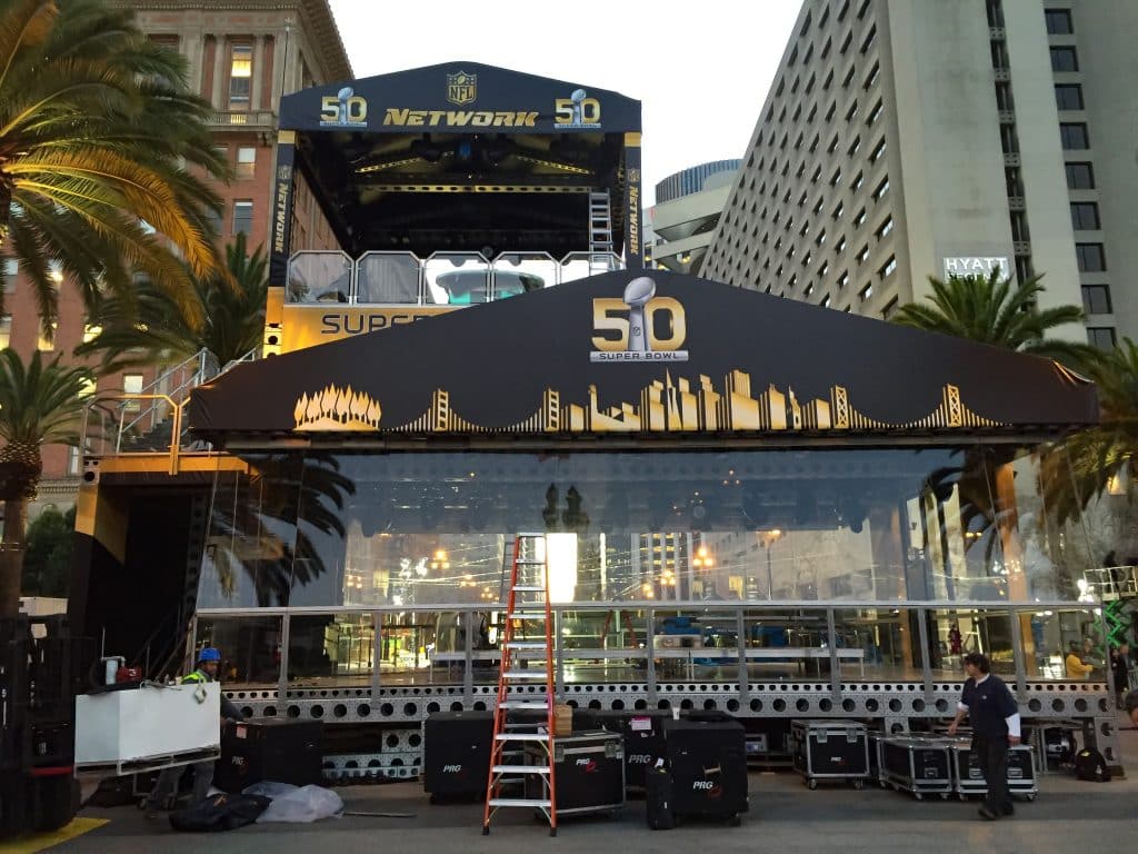 Front, ground level view of a multi-level structure covered in "NFL NETWORK" branded soft goods. Color scheme is black and gold. "50th Superbowl" can be seen on the front of the roof gable. behind the foreground structure is a taller one on the left side.