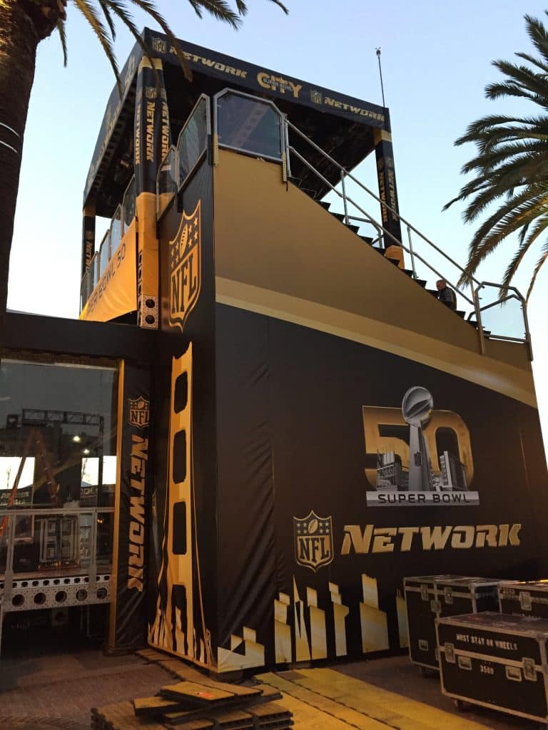 Side view of a multi-level structure covered in "NFL NETWORK" branded soft goods. Color scheme is black and gold. "50th Superbowl" can be seen on the side of the fabric. Sunset light shines on the left facing side of the structure and palm trees are on both sides.