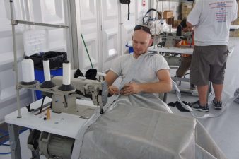 White male in white shirt is sitting at a white table in a white room, sewing a light gray soft goods on site at an event. More people are behind him also working.