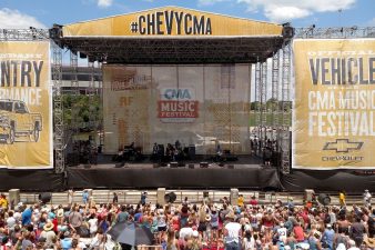 ChevyCMA stage with roof, backdrops and side wing softgood elements. The soft goods are all a yellow base with chevy logo and trucks images printed on them, along with "country performance", "official vehicle of the CMA music festival", and "chevy C M A". A crowd is in front of this daytime stage. Black scrim borders the bottom of the stage hiding the under structures.