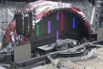 Custom keder panels over a truss arch that covers a stage, LED wall test screen is on, its day time and load in is still happening. POV is up audience left looking down from the upper level of a stadium