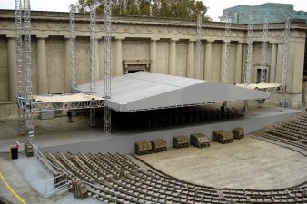 3/4 view audience view of a grey stage roof located outdoor inside a n off-white colored, greek-style amphitheater. The stage roof is lowered to about 8' above the stage decks. Semi-circe rows of chairs surround the front of the stage. There are no people in this photo.