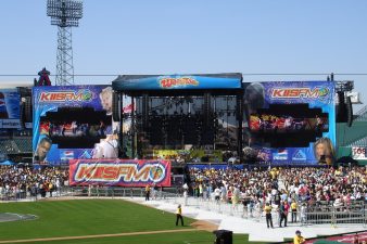Looking at a stage with colorful printed scrims on covering the side wings and black backdrop scrims hanging from the upstage to darken the back of the stage. The scrims have "Kiss FM 102.7" logos and images of pop and rap musicians all mashed together like the 80s.