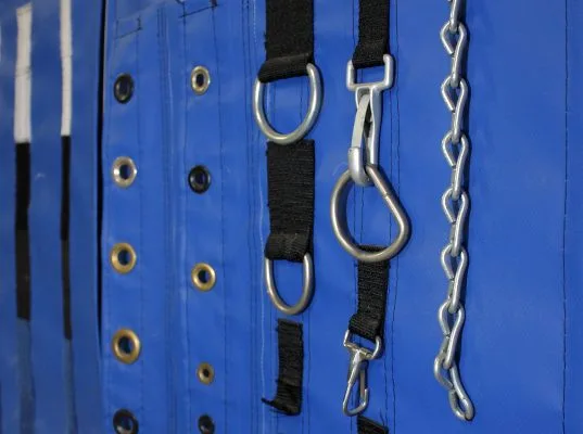 Cropped image: Various D-rings, grommets, and chains displayed on a blue vinyl wall. Accessories for attaching roof skins and side walls to stages and live event structures.